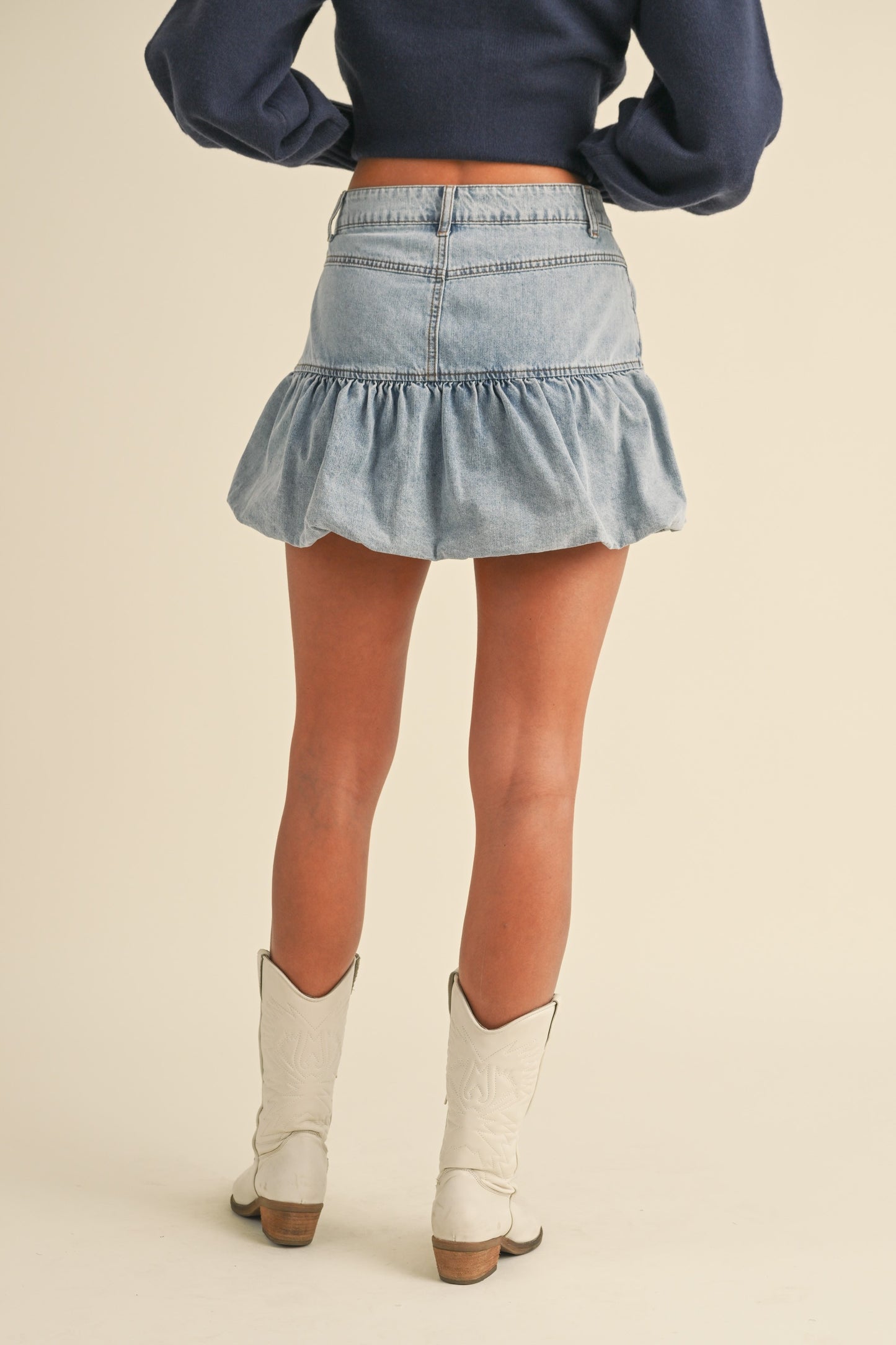 Back view of a model wearing a high-waisted denim bubble skirt with a ruffled hem. The light-wash jean skirt features structured seams and a fitted waist, creating a flattering silhouette. She pairs it with a navy cropped cardigan and white cowboy boots for a chic, modern western look.