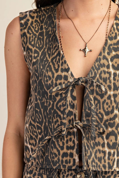 A close-up of a leopard print denim vest featuring two front tie closures. The fabric's bold pattern is highlighted, along with the intricate details of the ties. The model is also wearing a delicate gold necklace with a star-shaped pendant.